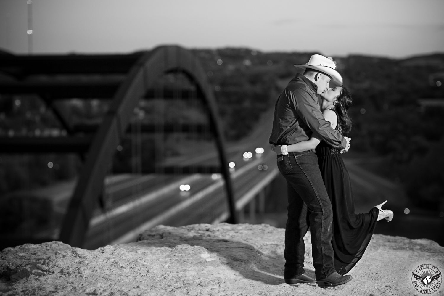 black and white image of a latino brunette girl with curly hair wearing a long black dress and tan heals kisses a guy in a cowboy hat and a dark shirt with blue jeans and boots on a cliff overlooking  the 360 bridge in the engagement image from austin  